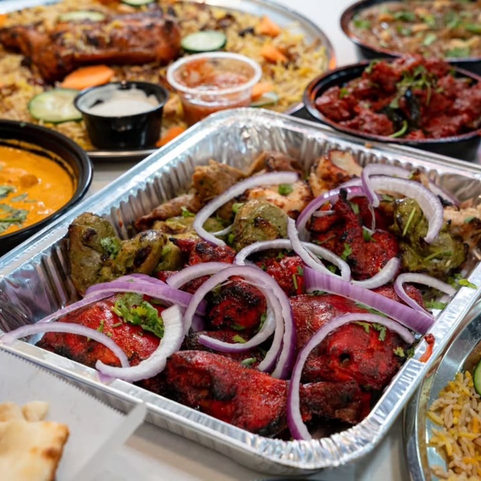 A foil tray of red and green tandoori chicken with sliced red onions, surrounded by other Indian dishes.