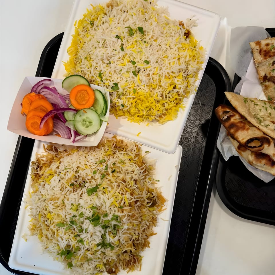 Two plates of Imperial Biryani with a side of sliced carrots, cucumbers, and red onions, plus naan.